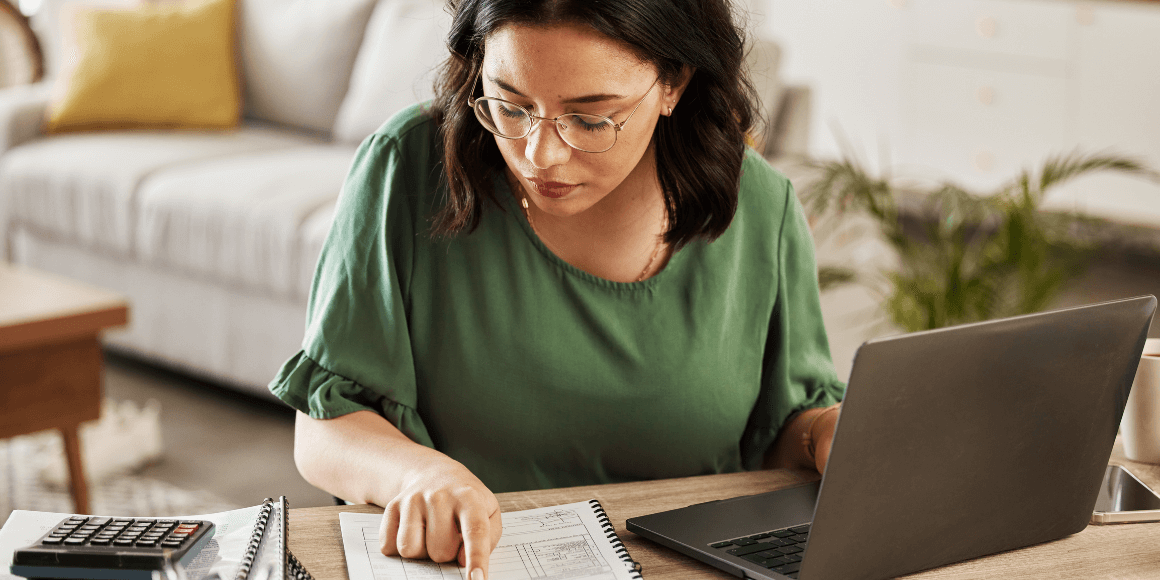 Woman sitting at her desk reviewing health savings documents and conducting research on her laptop.