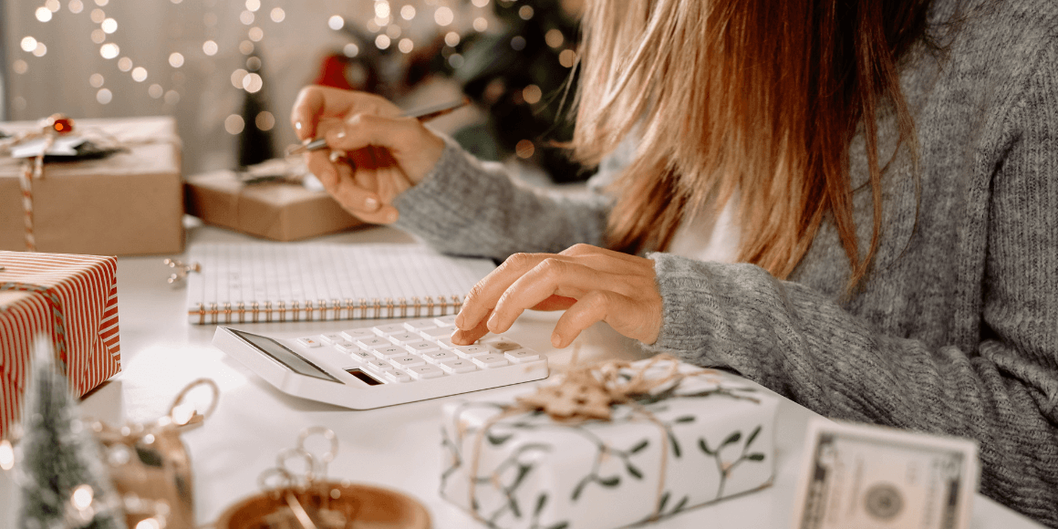 Person in a gray sweater using a calculator and writing in a notebook at a holiday decorated table with wrapped gifts and dollar bills nearby showing that they are working on a budget